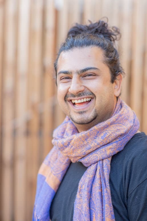 Portrait of Parham Farsi outdoors in soft natural light, wearing a cream tunic and an earthy patterned scarf, against a softly blurred green woodland background